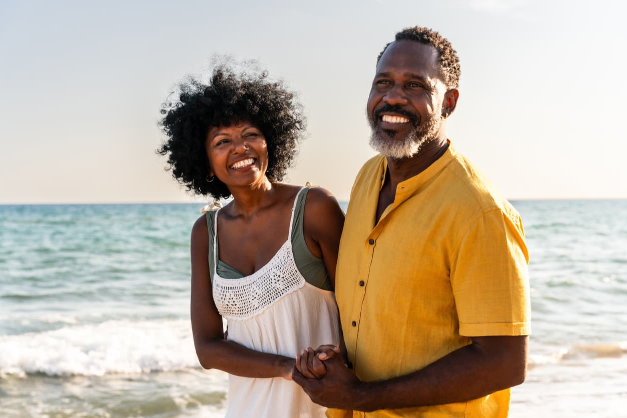 A couple smiles warmly while standing close on a sunny beach. The woman wears a white dress, and the man wears a yellow shirt. The ocean waves gently roll behind them.