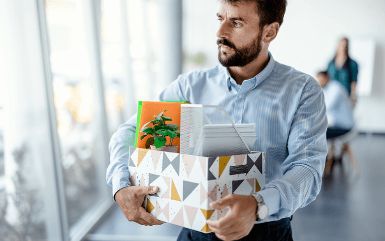 A man with a serious expression holds a decorative box containing office supplies and a small plant, walking in a bright office with blurred colleagues in the background.