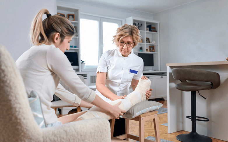 A nurse tends to a woman's bandaged leg in a cozy home setting. The woman appears concerned as the nurse gently examines her injury.