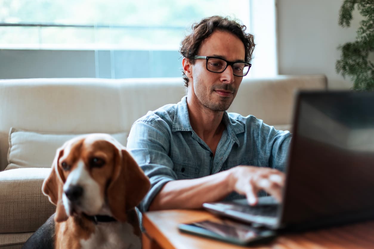 A man is focused on his laptop while sitting on a couch. He wears glasses and a casual shirt. Beside him, a calm beagle looks towards the camera.