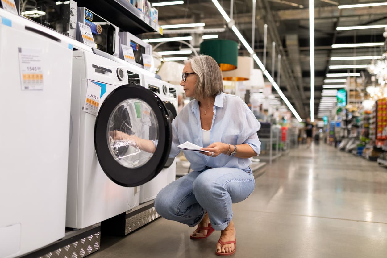 A woman with silver hair and glasses examines a front-loading washing machine in a large retail store. She holds product information while crouching to inspect the appliance, thoughtfully considering a major purchase.