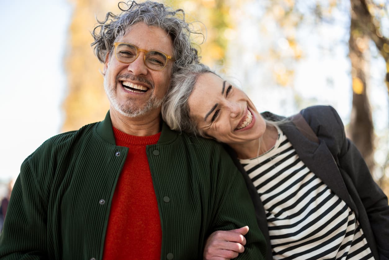 Smiling couple in casual attire embrace outdoors, with autumn trees in soft focus. They display warmth and happiness, leaning close together.