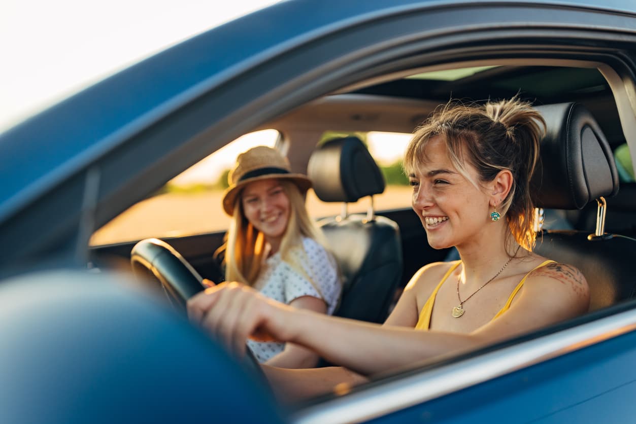 Two women in a car, one driving and smiling, the other in a sun hat in the passenger seat. They appear happy, with a sunlit background.