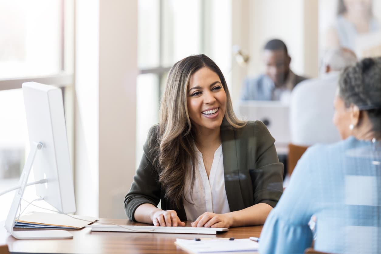 A smiling young woman with long brown hair in an olive blazer provides friendly customer service across a wooden desk. She faces an older woman with short gray hair in a bright, modern office with large windows and natural light.