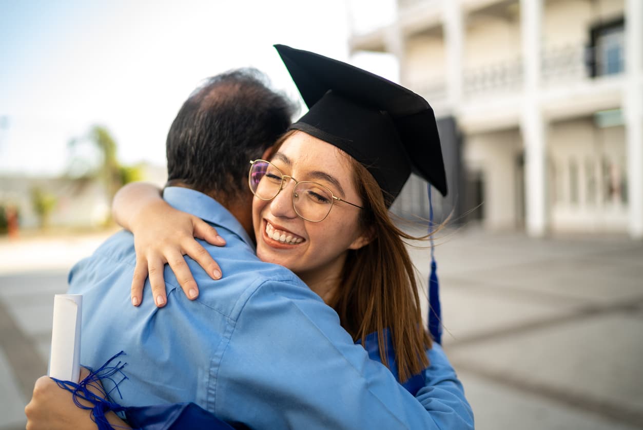 A young woman graduate wearing a black cap and blue gown joyfully embraces a family member outdoors. She holds a rolled diploma tied with a blue ribbon, her eyes crinkled with genuine happiness as she celebrates this milestone moment.