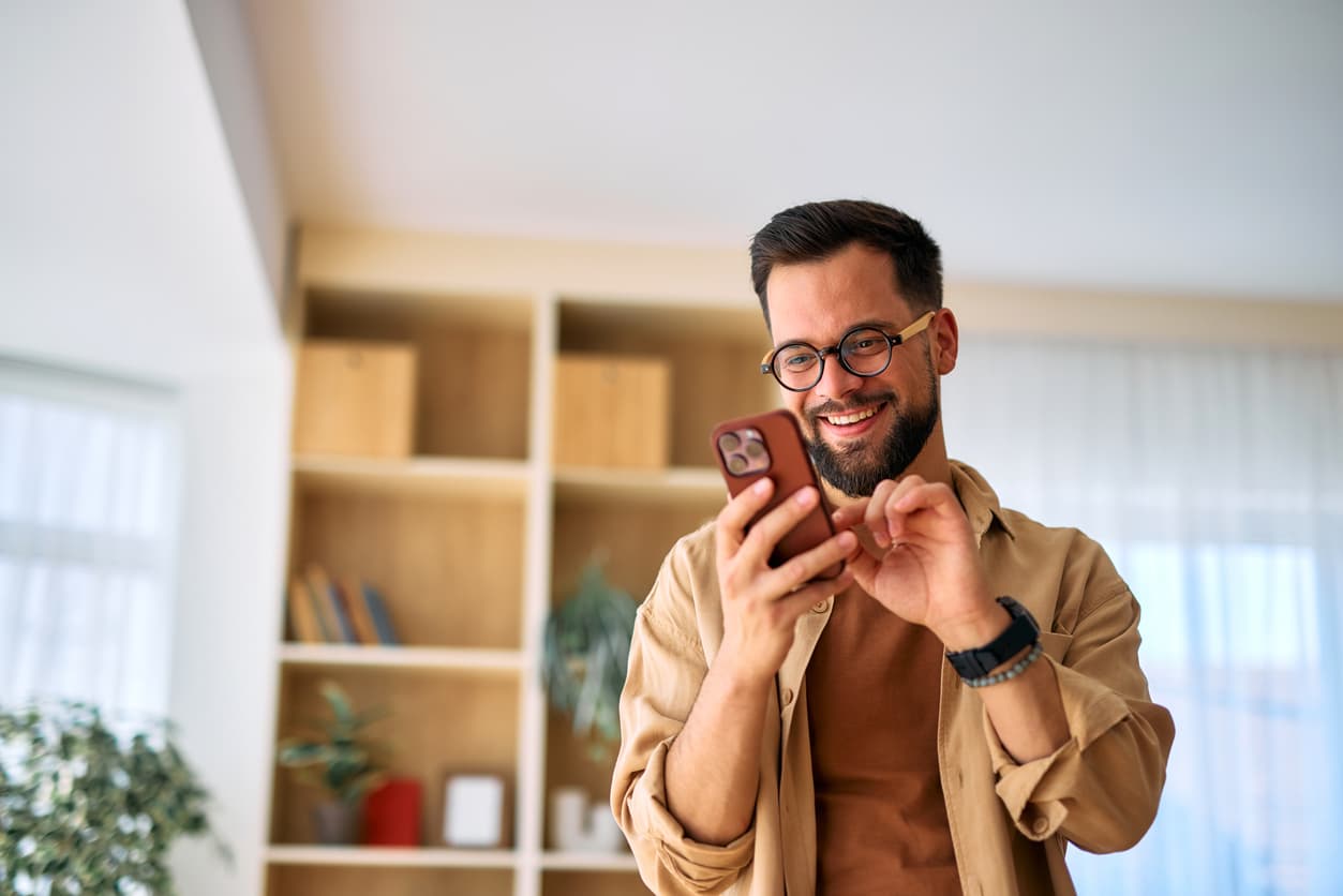 Smiling man with glasses looks at smartphone indoors, wearing a beige shirt. Light wood shelves and a plant in the background convey a cheerful mood.