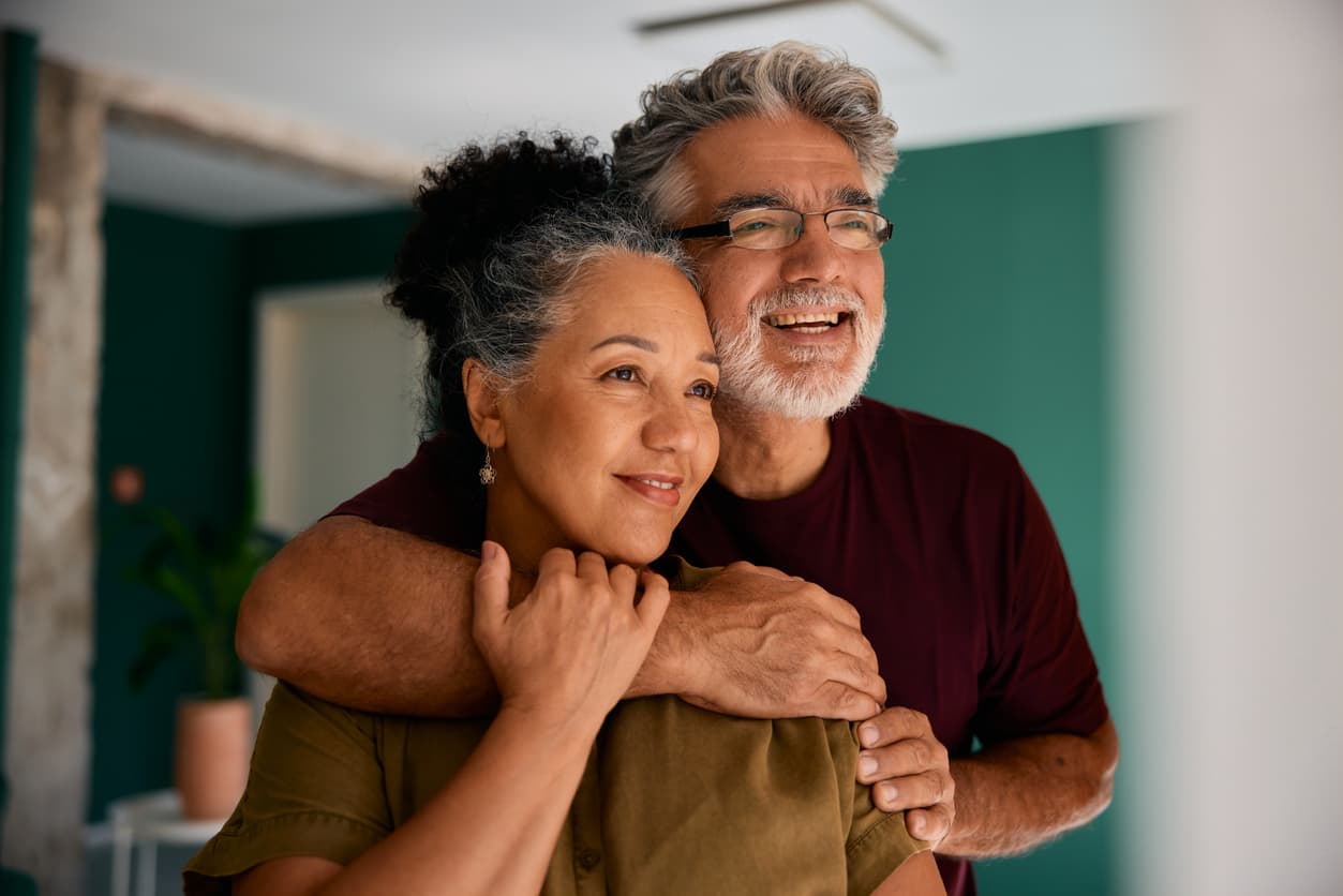 A loving couple in their 50s-60s shares an affectionate embrace in their modern home. The gray-haired man wraps his arms around the woman from behind, both laughing joyfully. A teal accent wall and natural lighting convey comfort and partnership.