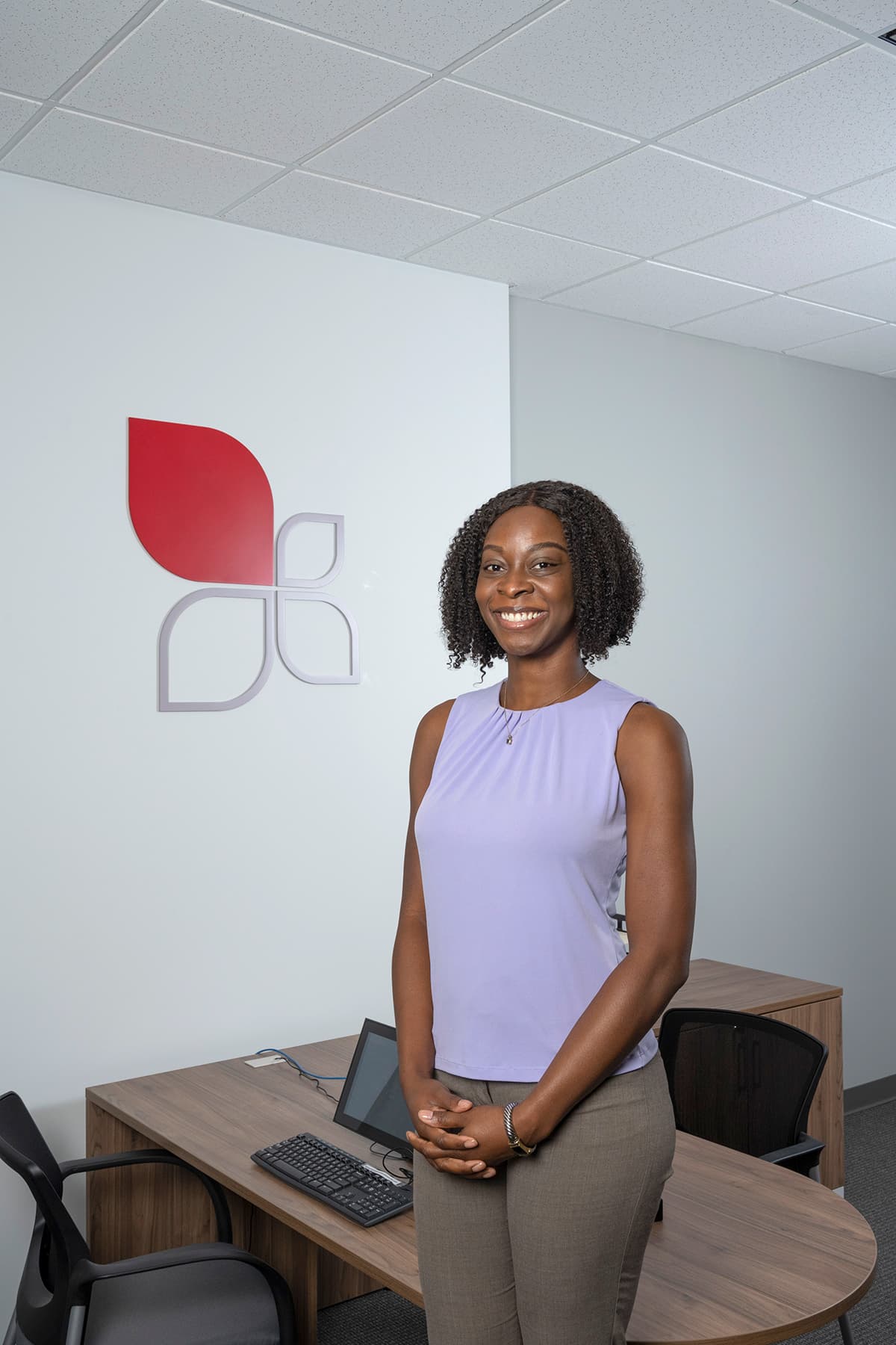 A confident young Black woman with natural curly hair stands behind a wooden desk in a modern office, smiling warmly. She wears a lavender sleeveless blouse and gray pants. The Lendmark corporate logo is visible on the white wall behind her.