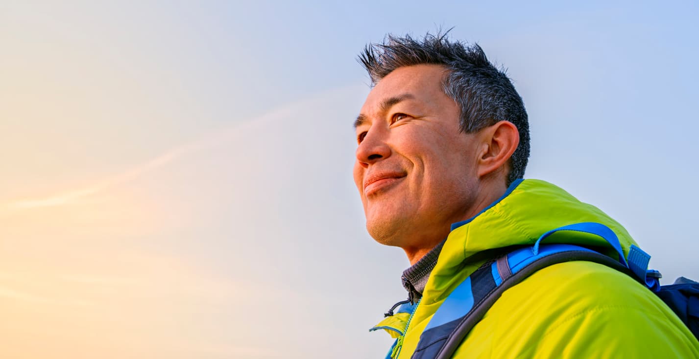 A middle-aged man with short dark hair smiles contentedly while hiking outdoors. He wears a bright lime green jacket with blue backpack straps visible. The golden hour sky creates a warm, uplifting mood of adventure and optimism.
