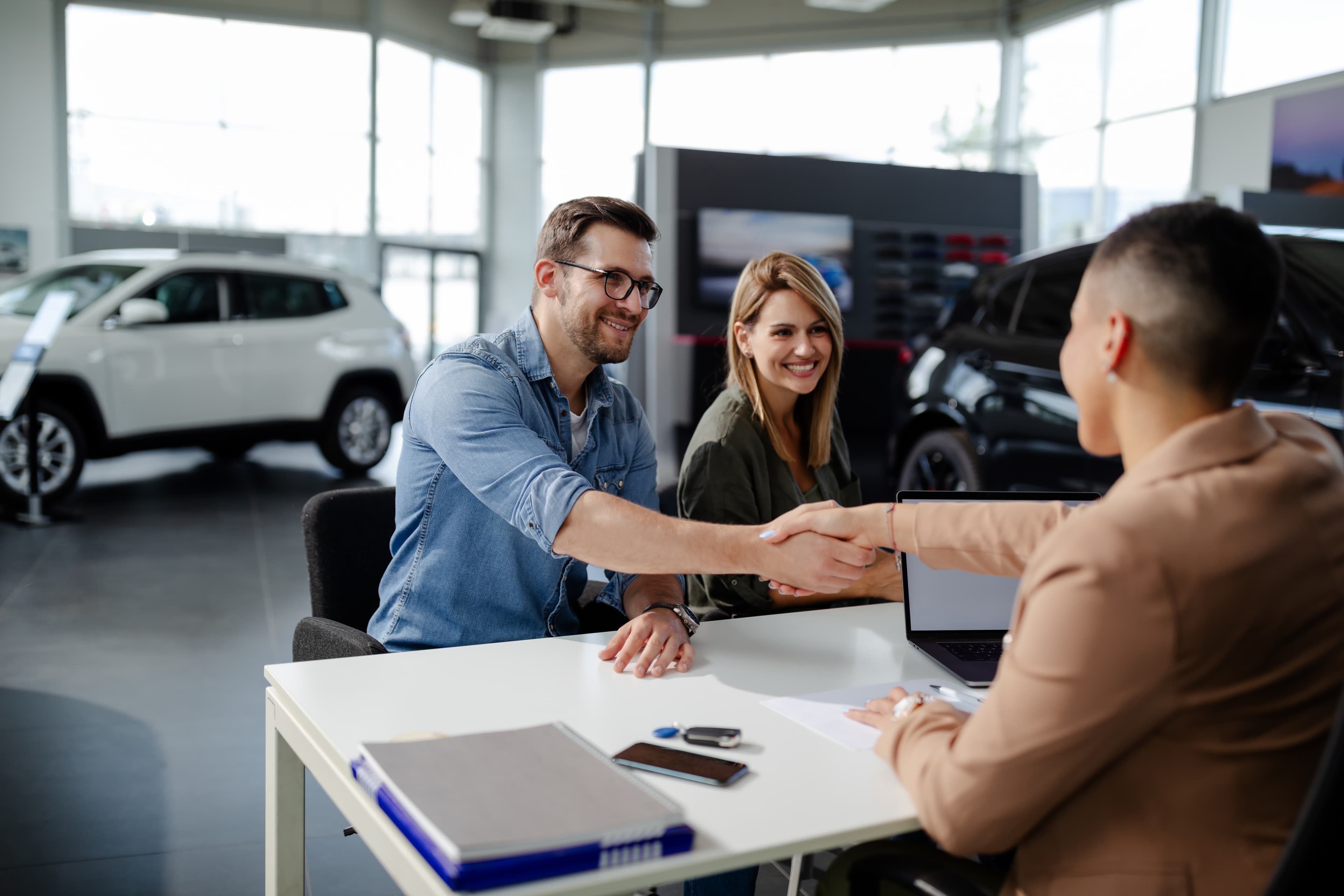 A smiling couple shakes hands with a salesperson at a table in a bright car dealership, with two cars visible in the background. The mood is positive.