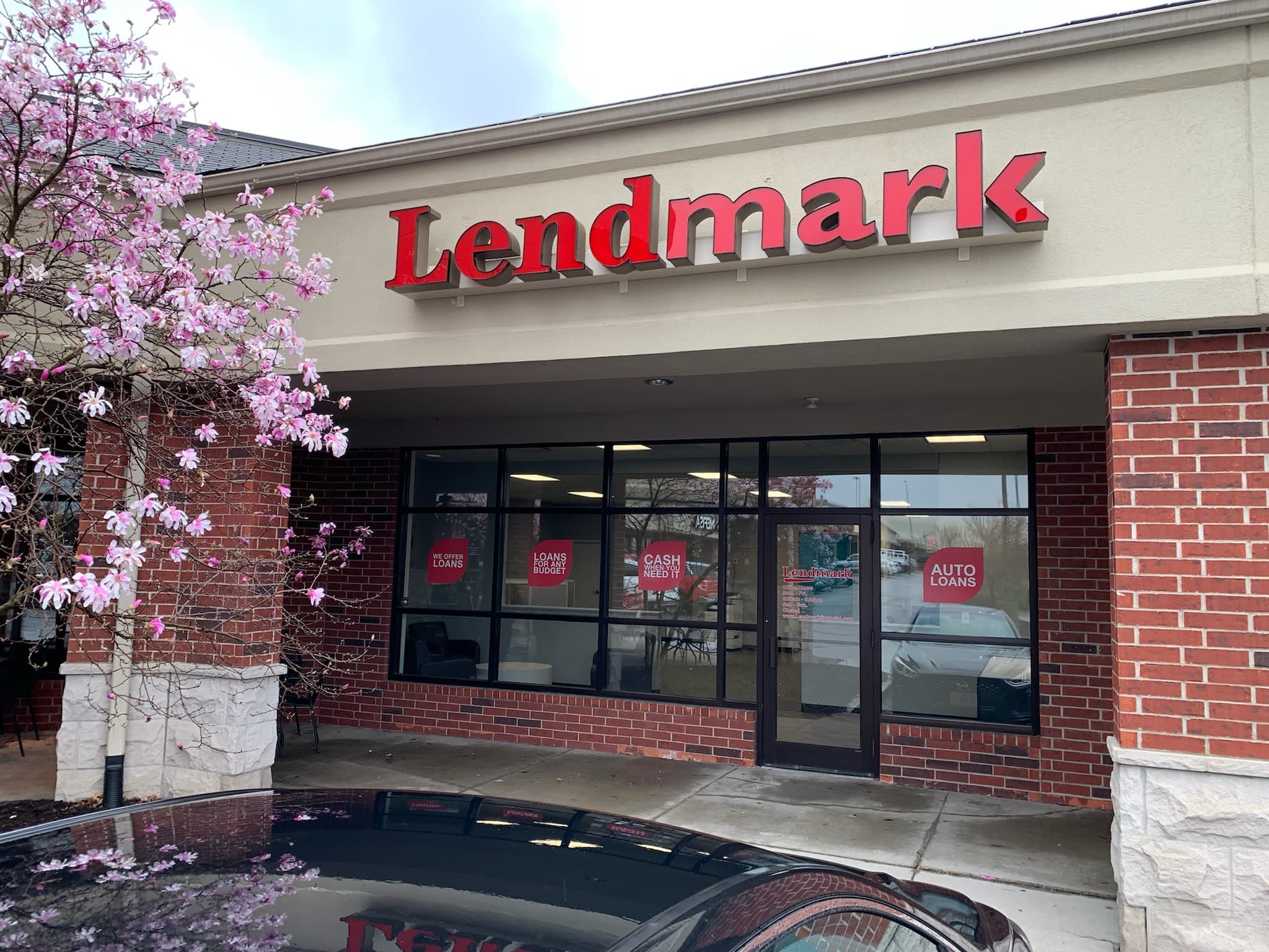 Lendmark Financial Services storefront on a spring day with red brick walls and beige stucco displaying the company name. Large windows show loan promotional signs, and a pink magnolia tree in bloom frames the welcoming entrance.