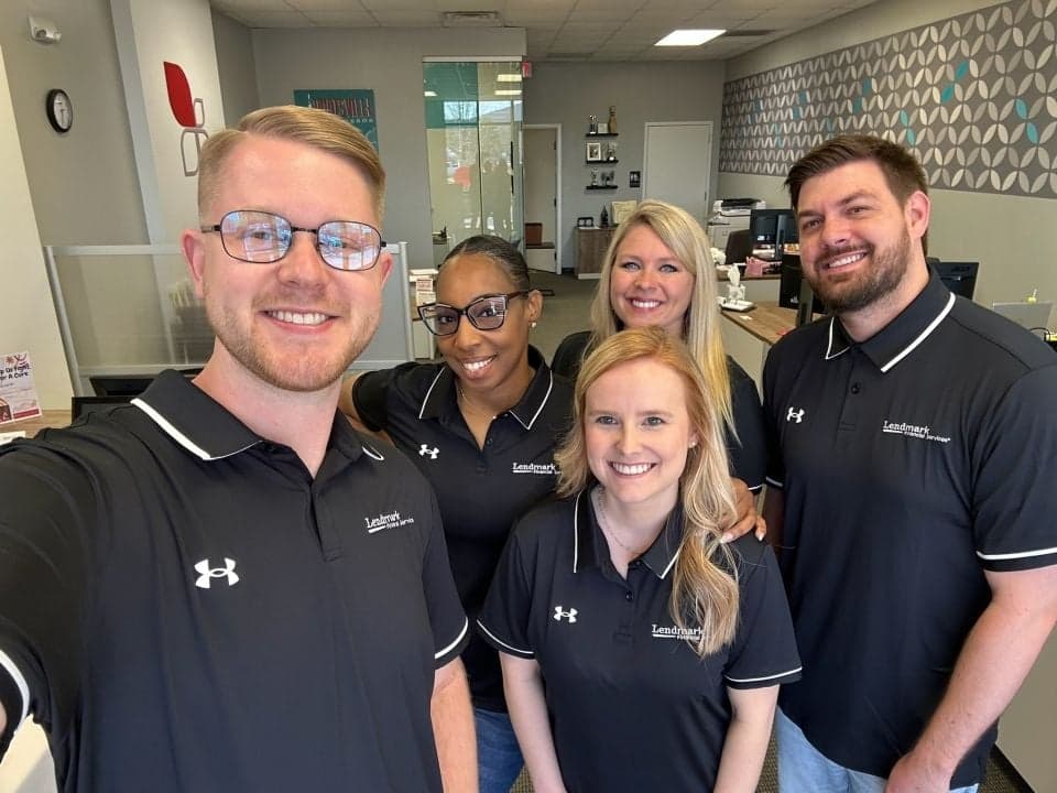 Five Lendmark employees pose for a team selfie in navy blue polo shirts. Behind them is a decorative wall with a geometric leaf pattern and a "Huntsville" sign. The team displays warm smiles, conveying workplace camaraderie.
