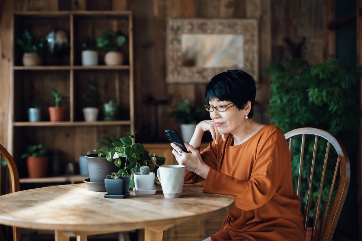 A woman with short black hair and glasses sits at a table with plants and a mug, looking at her smartphone.